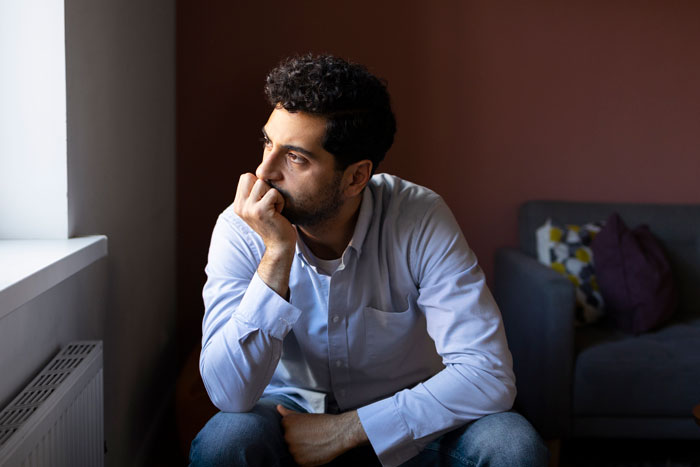 Man looking thoughtful and worried by a window, representing a husband who feels sick after realizing their son’s namesake. - 4