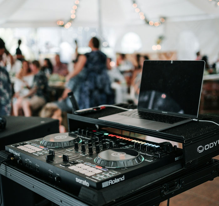 DJ equipment and laptop set up at a wedding reception with guests in the background, capturing the event atmosphere. - 10