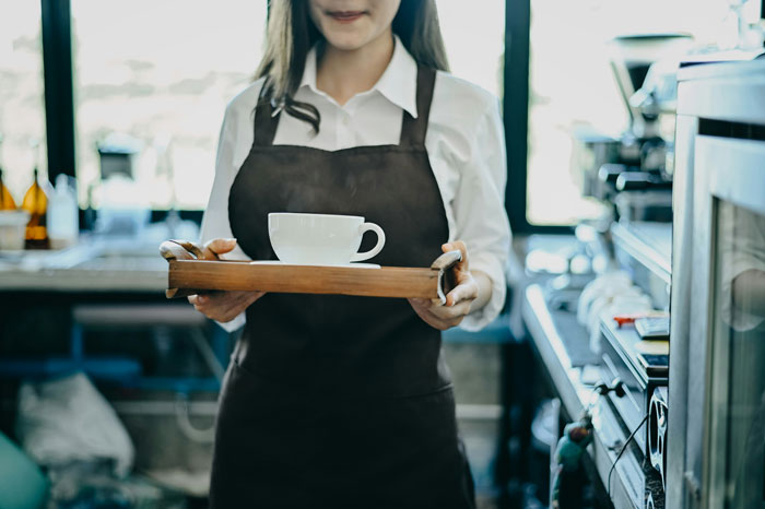 Woman wearing apron holding tray with cup in cafe, representing women sharing inappropriate comments heard in public spaces.