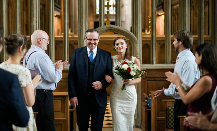 Bride walking down the aisle with her father at a wedding, guests applauding in a historic church setting. - 41