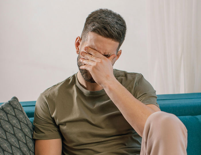 Man sitting on couch, covering his face with hand, appearing upset or stressed, representing relationship struggles.