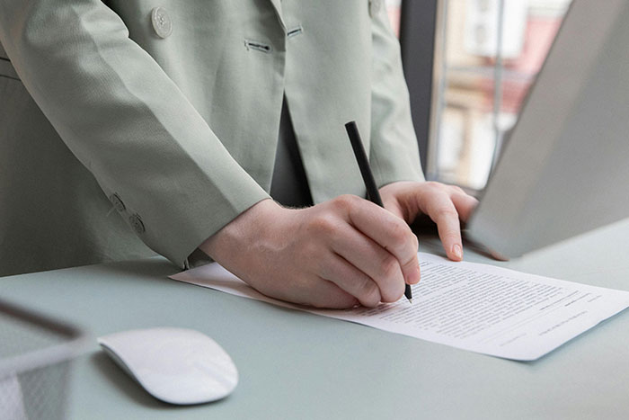 Person in a green suit jacket writing on a document with a pen, illustrating microfeminisms and social commentary.