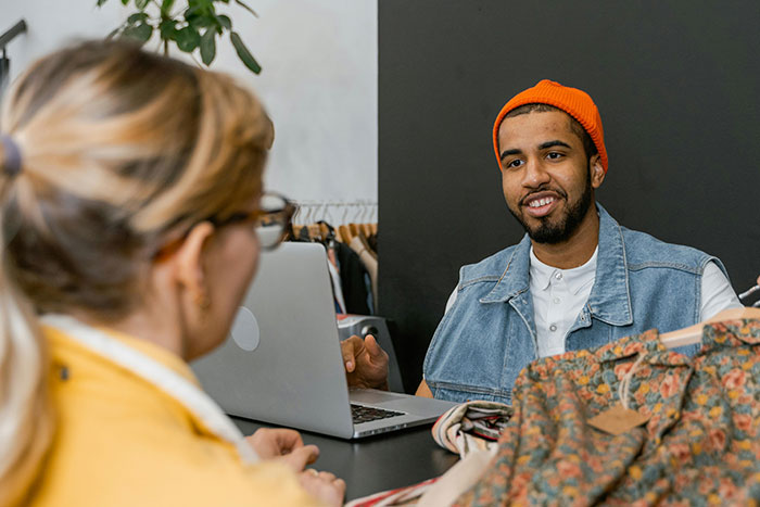 Man wearing orange beanie and denim vest, sitting at a table with laptop while talking to a woman in a yellow jacket.