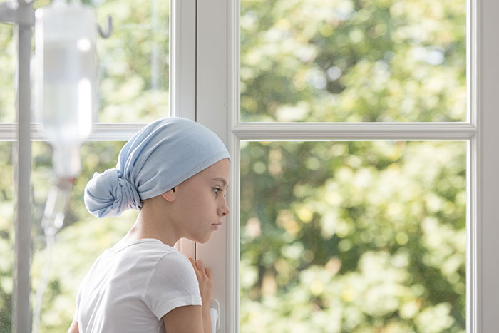 Teen girl with headscarf looking out window, symbolizing family upset over 17YO not shaving head to support 7YO with cancer.
