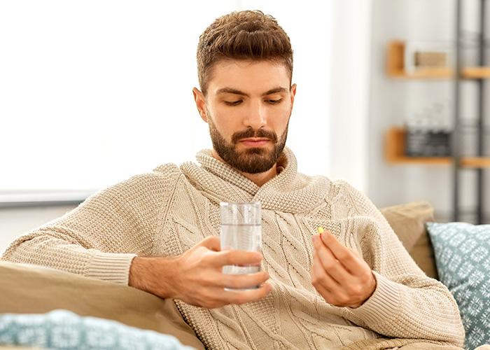 Young man in a beige sweater sitting on a couch, holding a pill and glass of water, showing facts people are tired of explaining.