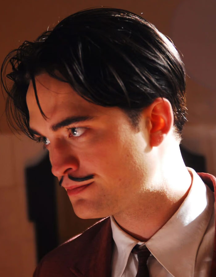Young man with dark hair and mustache in a brown suit, portraying a scene related to Robert Pattinson Twilight intimate scene.