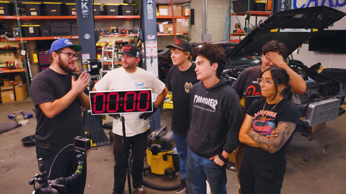 A group of people share unsubscribed YouTubers opinions in a garage workshop with cars and tools around.