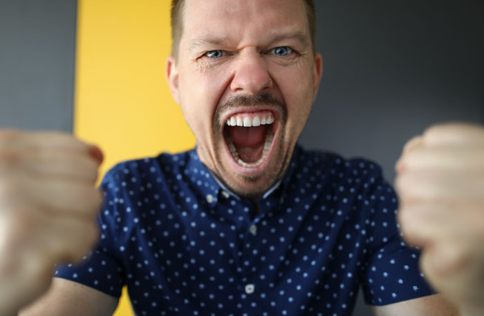 Man in a blue patterned shirt expressing excitement with clenched fists and an open mouth in front of a yellow and gray background.
