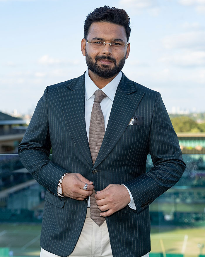 Man in pinstripe suit and glasses posing outdoors with tennis courts in the background at a Wimbledon event.