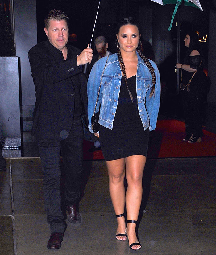 Demi Lovato walking with bodyguard holding umbrella over her outside at night in a black dress and denim jacket. Demi Lovato walking with bodyguard holding umbrella over her outside at night in a black dress and denim jacket.