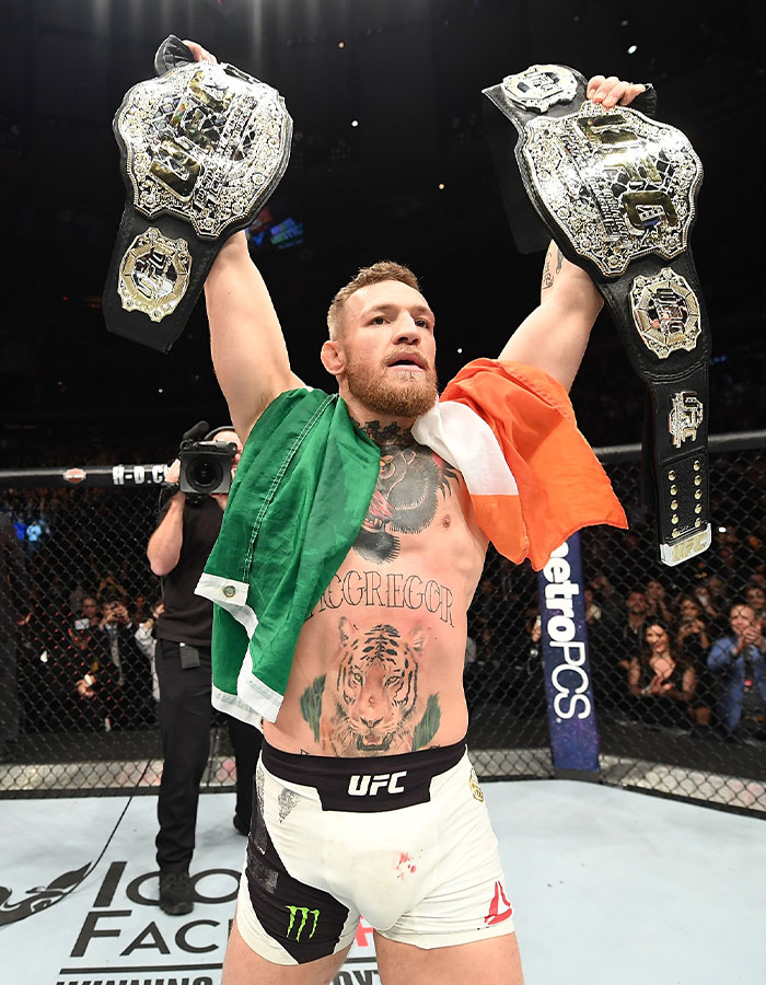 Conor McGregor celebrating victory in UFC octagon, holding two championship belts with Irish flag draped over shoulders.