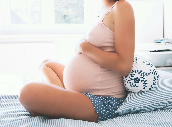 Pregnant woman sitting on bed in a bright room, highlighting a tense or emotional moment related to messed up school incidents. - 18