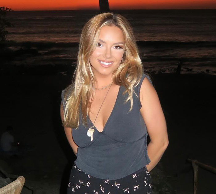 Woman who recorded CEO cheating on Coldplay kiss cam smiling at sunset near the beach wearing dark top and floral skirt.