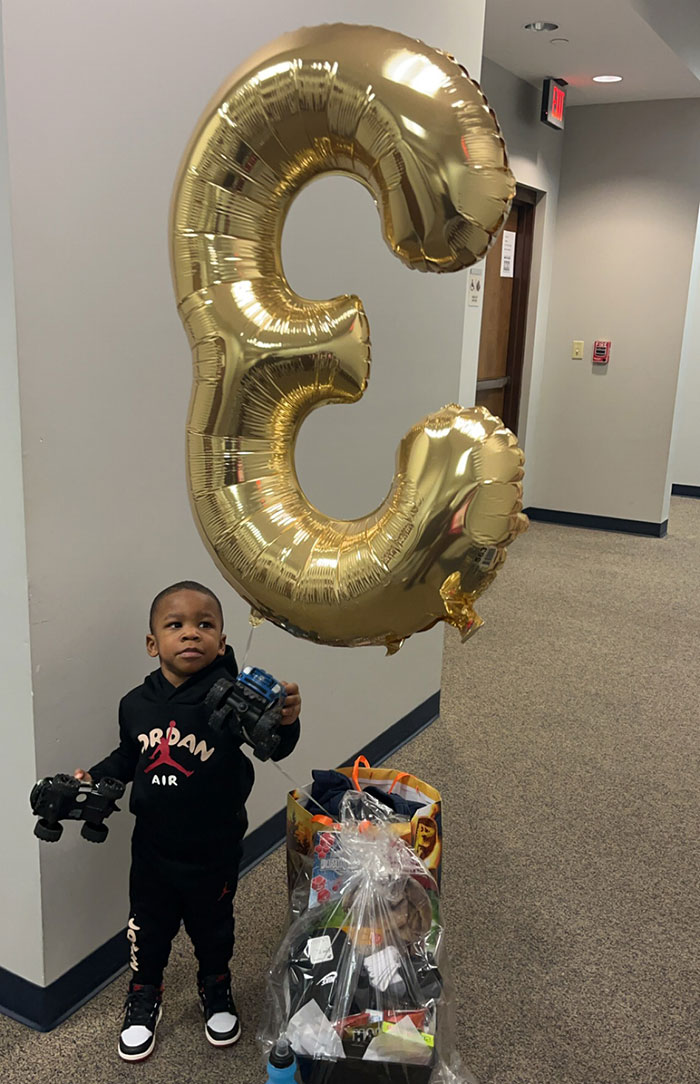 3-year-old boy in temporary foster care holding toy cars next to large gold number three balloon in hallway setting - 1