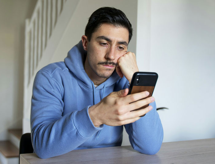 Man in a blue hoodie looking worriedly at his phone, representing jealous wife spying husband work concerns.