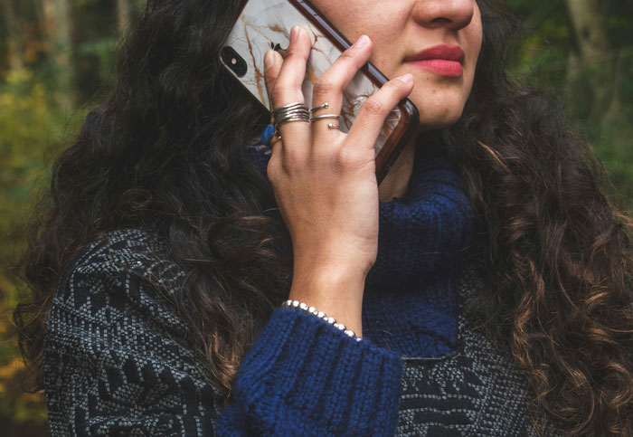 Woman with curly hair wearing rings and a sweater talking on the phone in an outdoor forest setting, tourist concept. - 10