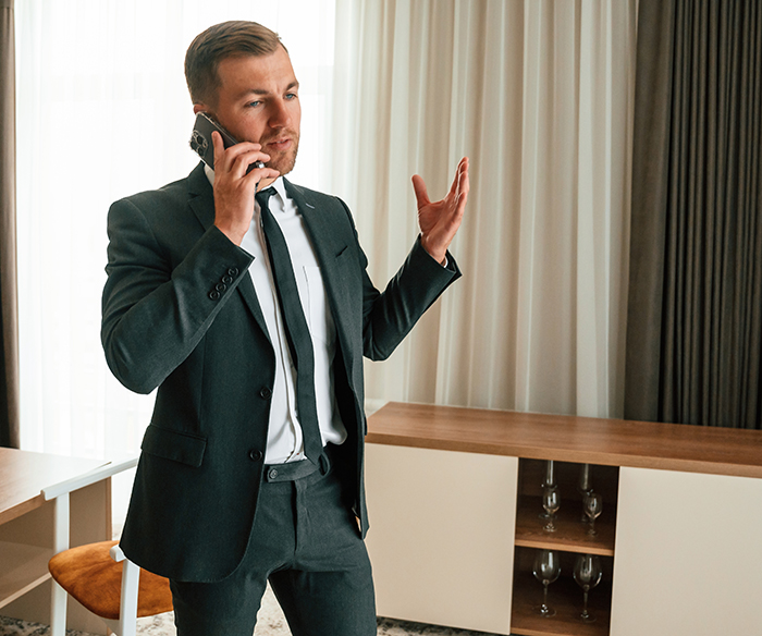 Man in a suit talking on phone in a hotel room, possibly a guest or hotel employee handling a situation.