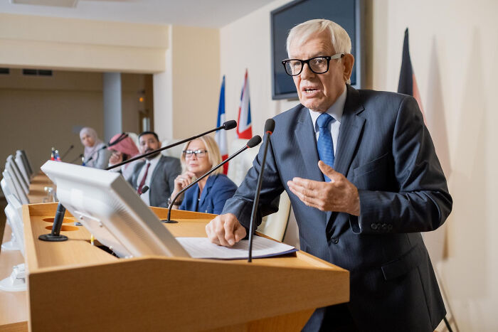 Older man in suit speaking at podium during a conference while others listen, illustrating normal people's habits discussion. - 10