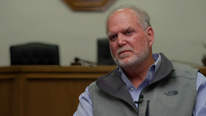 Middle-aged man with gray hair and beard, wearing a North Face vest, discussing consequences of stupidity and job loss.