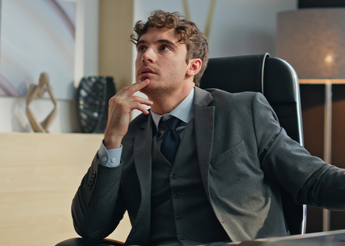 Young businessman in a suit sitting in an office chair, thoughtfully considering ridiculous complaints from employees.
