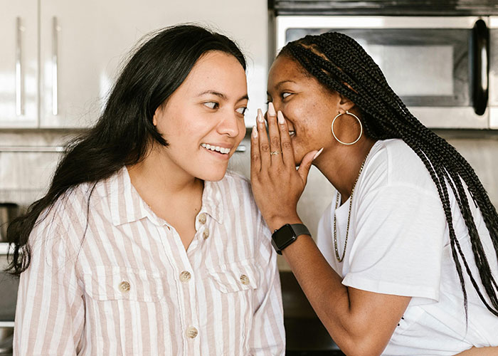 Two women whispering and smiling in a casual setting, illustrating workplace communication and micromanagement dynamics.
