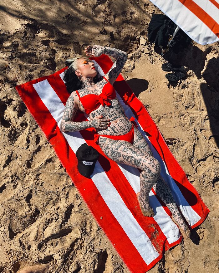 Woman with extensive body suit tattoos sunbathing on a red and white striped towel at the beach.