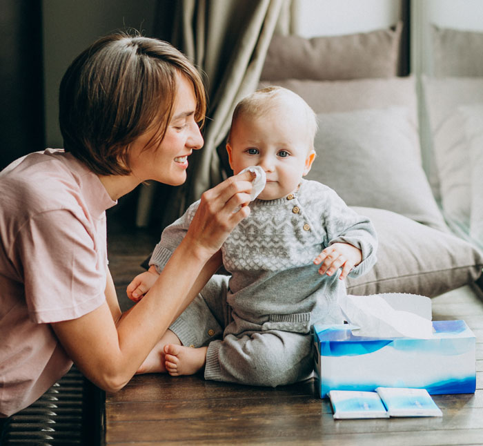 Nanny caring for baby indoors, gently wiping the child's face while sitting next to tissues on a wooden surface. - 12