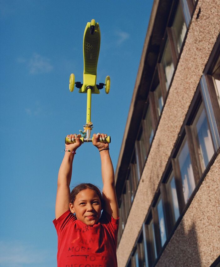 Child holding a green scooter above head outdoors in London, captured in honest street photography by Nico Froehlich.