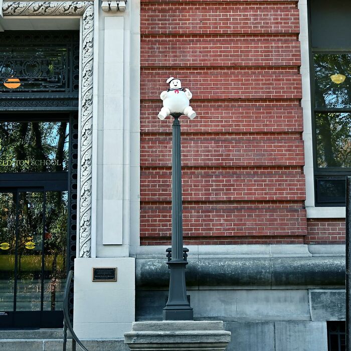 Street art interpretation showing a Stay Puft Marshmallow Man sculpture on a lamppost against a brick wall.