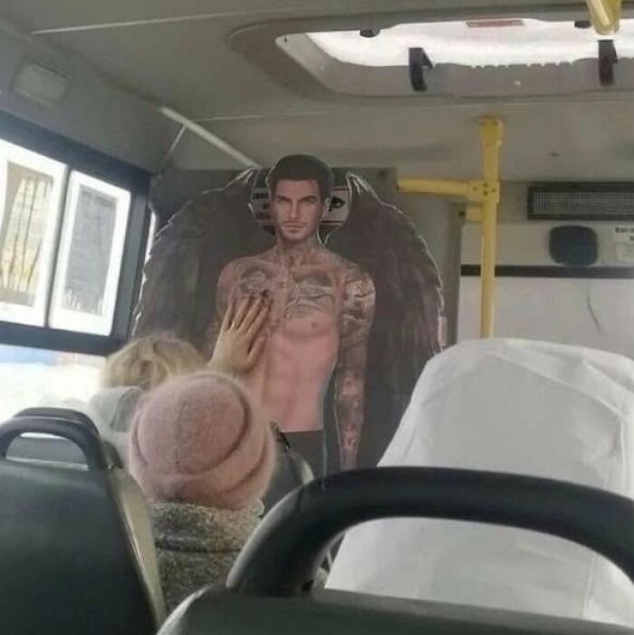 Person touching a cardboard cutout of a tattooed shirtless man with wings inside a crowded bus, combining trash and culture humor.