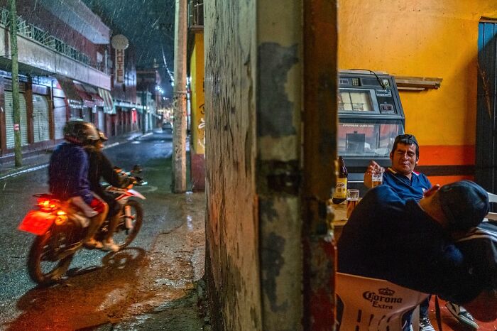 Two men enjoying drinks inside a small bar while a motorbike passes by on a rainy street, honest street moments captured.