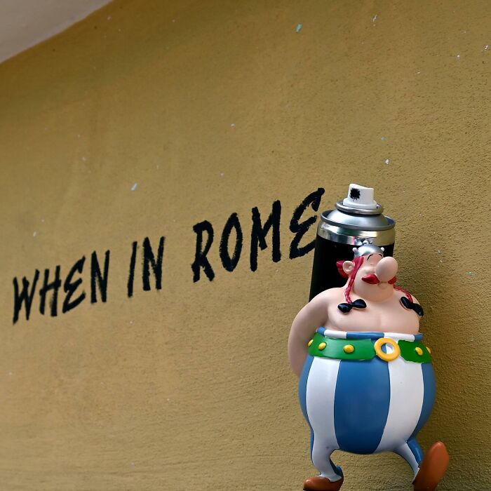 Street art featuring a small figurine on a ledge and the phrase when in Rome on a yellow public space wall.