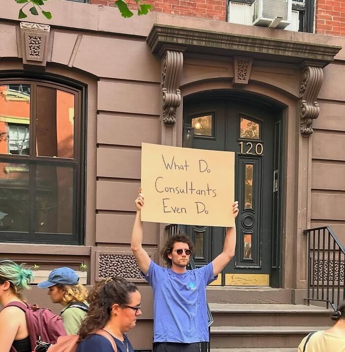 Man holding a Dude With The Sign cardboard sign that reads What Do Consultants Even Do on a city sidewalk.