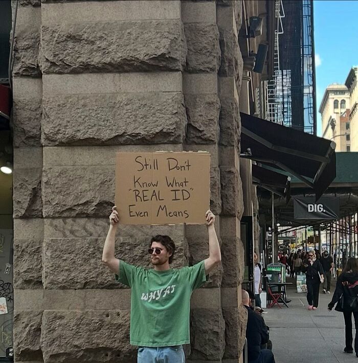 Dude With The Sign holding a cardboard sign about real ID, standing on a city street with pedestrians and buildings.