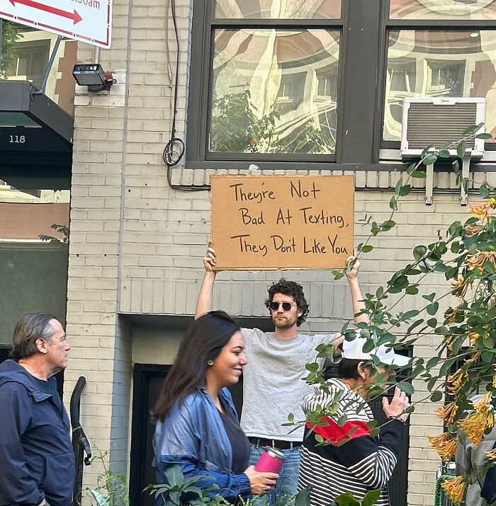 Man in sunglasses holding a Dude With The Sign cardboard with a humorous texting message on a city street.
