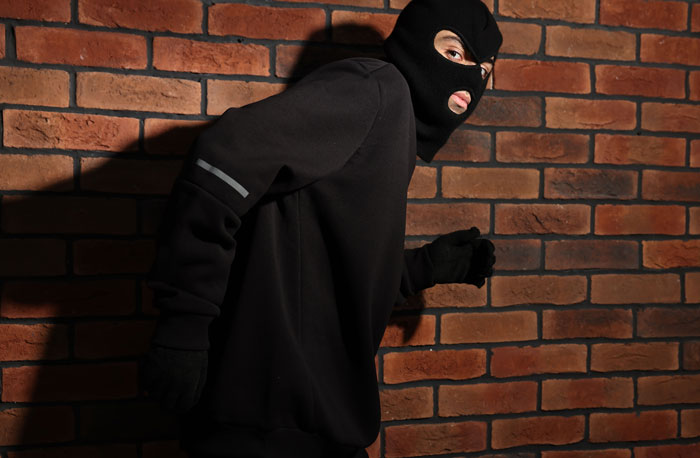 Person wearing a black balaclava and gloves, sneaking against a brick wall, illustrating tips to stay safe and alive.