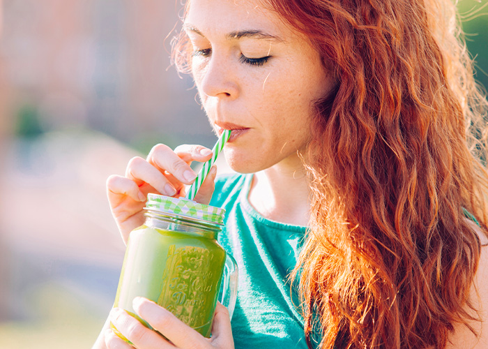 Young woman with red hair sipping a green smoothie outdoors, illustrating facts people are tired of explaining.