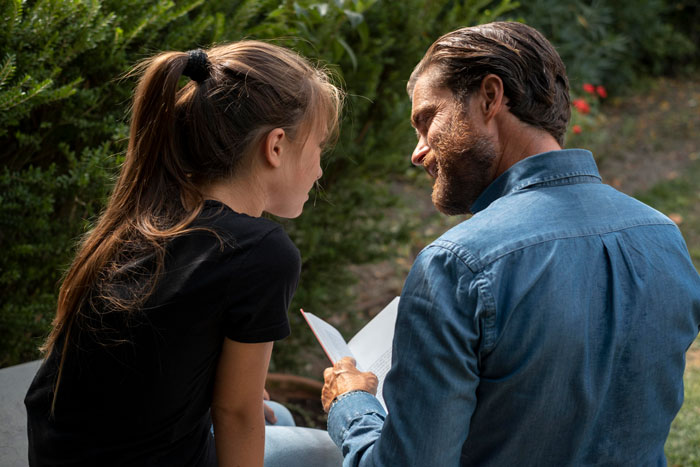 Father and daughter sitting outdoors, sharing a moment while reading a book during a menstruation celebration event. - 1