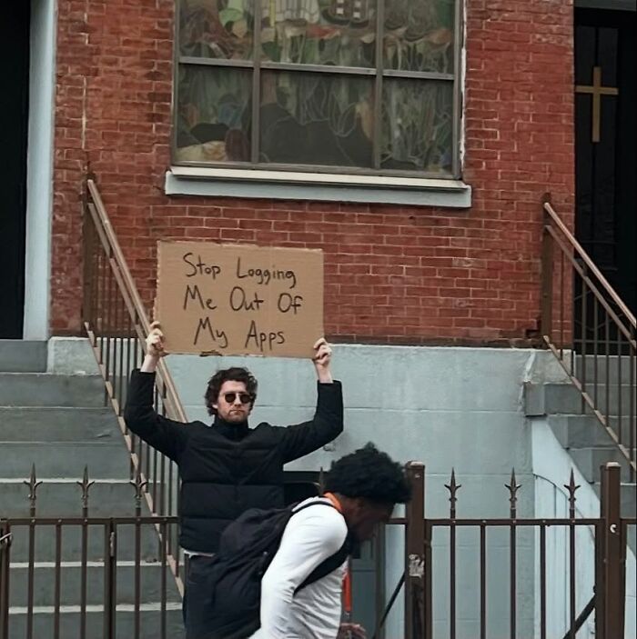 Man holding a Dude With The Sign cardboard sign saying stop logging me out of my apps on a city street.