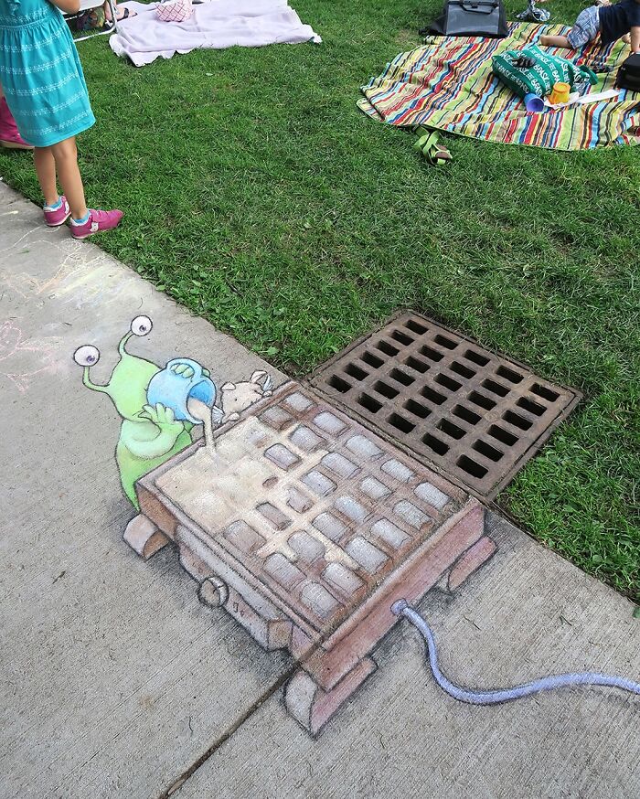 Sidewalk chalk art by David Zinn showing a green alien pulling a winged pig with a rope on the street.