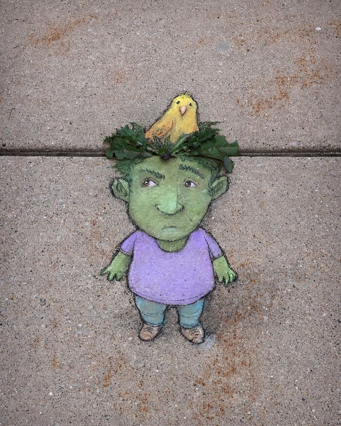Chalk drawing by David Zinn on sidewalk of a small green character with a bird on its head surrounded by leaves.