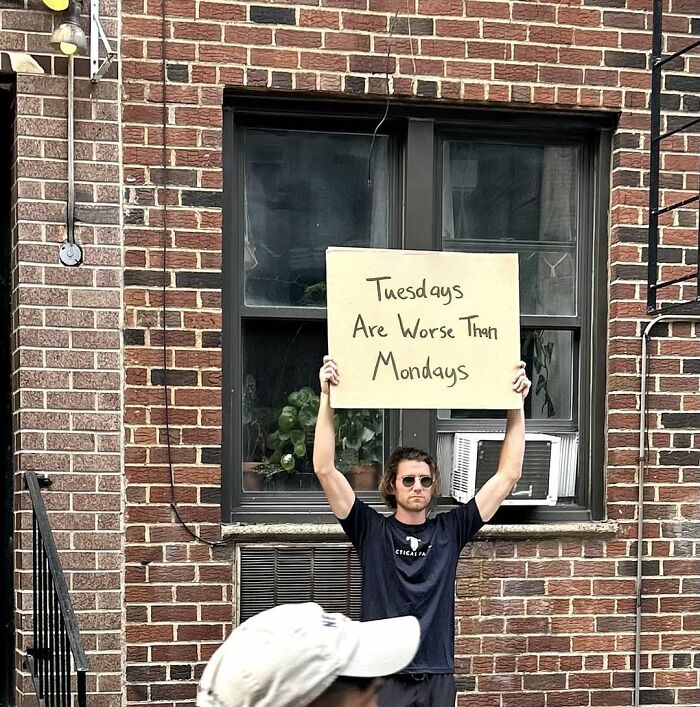 Man from Dude With The Sign holding a cardboard sign saying Tuesdays are worse than Mondays outside a brick building.