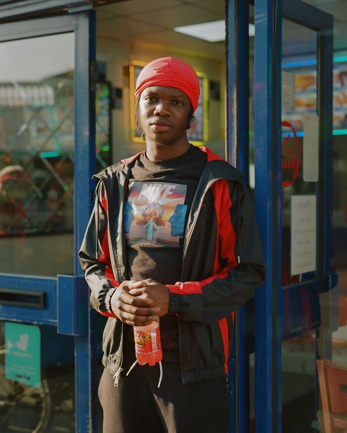 Young man wearing a red headscarf and black jacket holding a drink in an honest photo of London streets.
