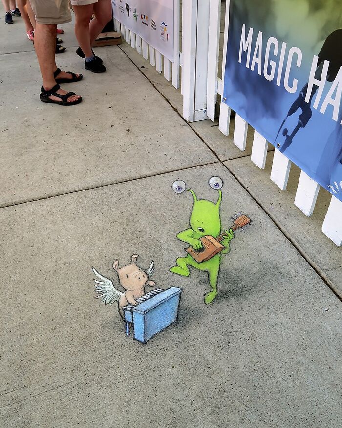 Sidewalk chalk art by David Zinn showing a green alien pulling a winged pig with a rope on the street.