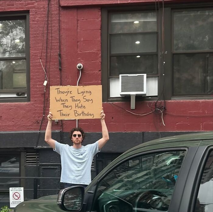 Man holding a sign that says they’re lying when they say they hate their birthday, Dude With The Sign in an urban setting.