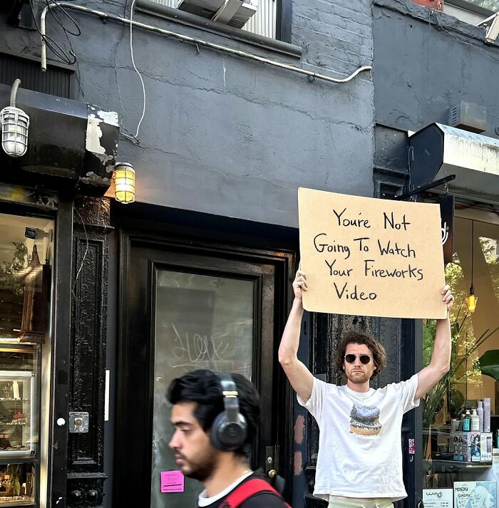 Man holding Dude With The Sign cardboard message about not watching fireworks video on city street background.