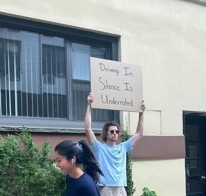 Man holding a sign outdoors with the message driving in silence is underrated, representing dude with the sign humor.