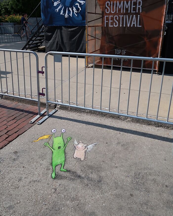 Sidewalk chalk art by David Zinn showing a green alien pulling a winged pig with a rope on the street.
