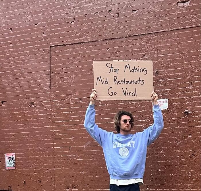 Man wearing sunglasses holding a Dude With The Sign cardboard sign against a red brick wall outdoors.