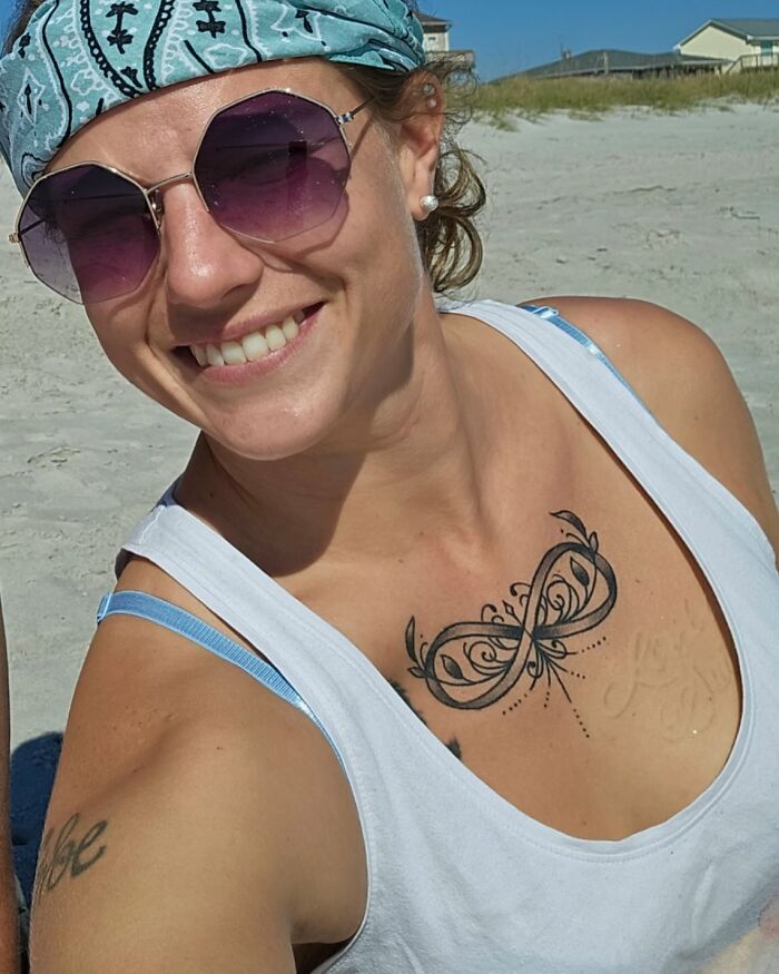 Smiling woman with sunglasses and a headscarf, showing an intricate infinity tattoo on her chest at a sunny beach. - 8